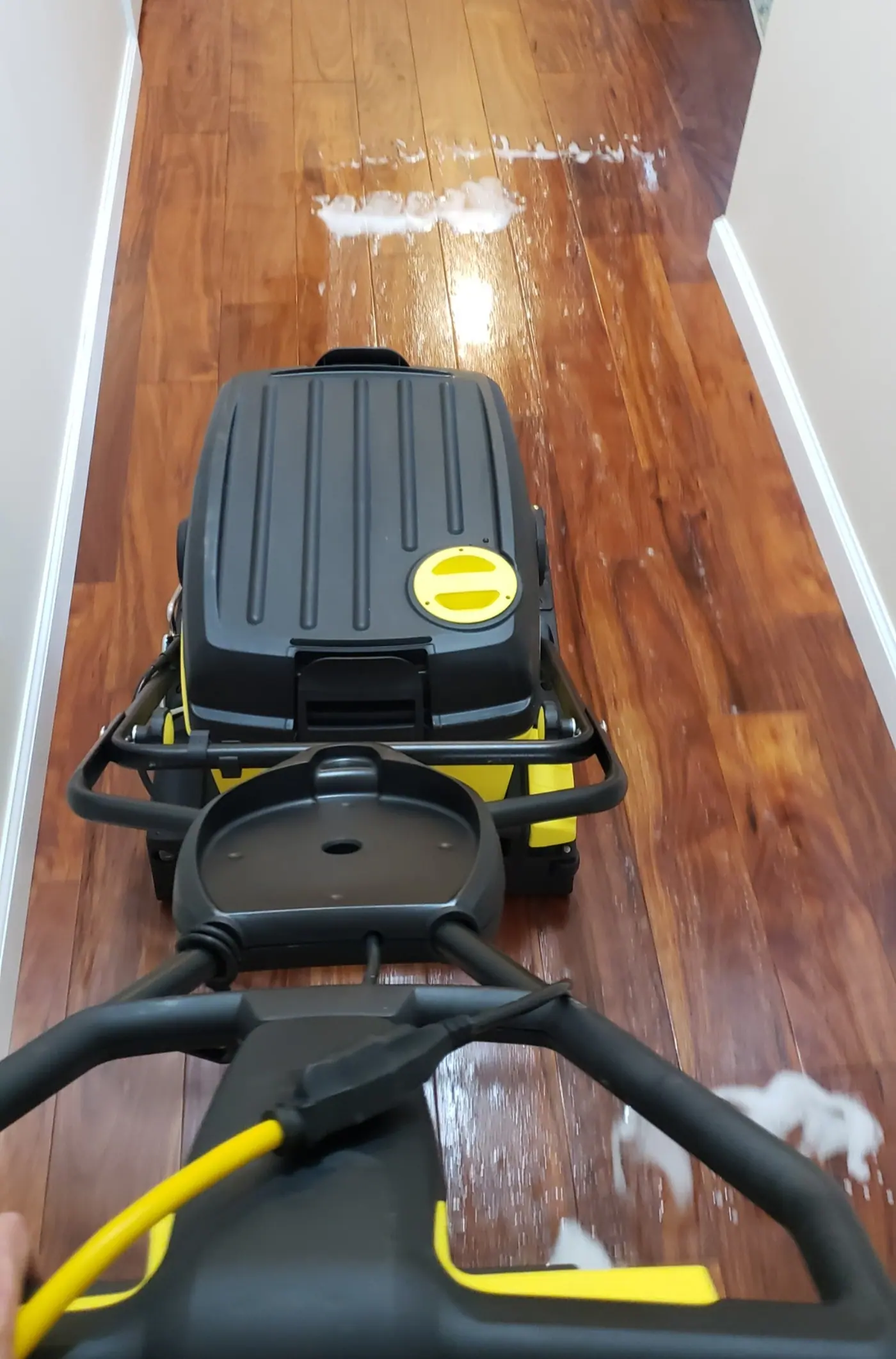 A floor scrubber cleaning a hardwood hallway, with a vacuum hose along the wall.