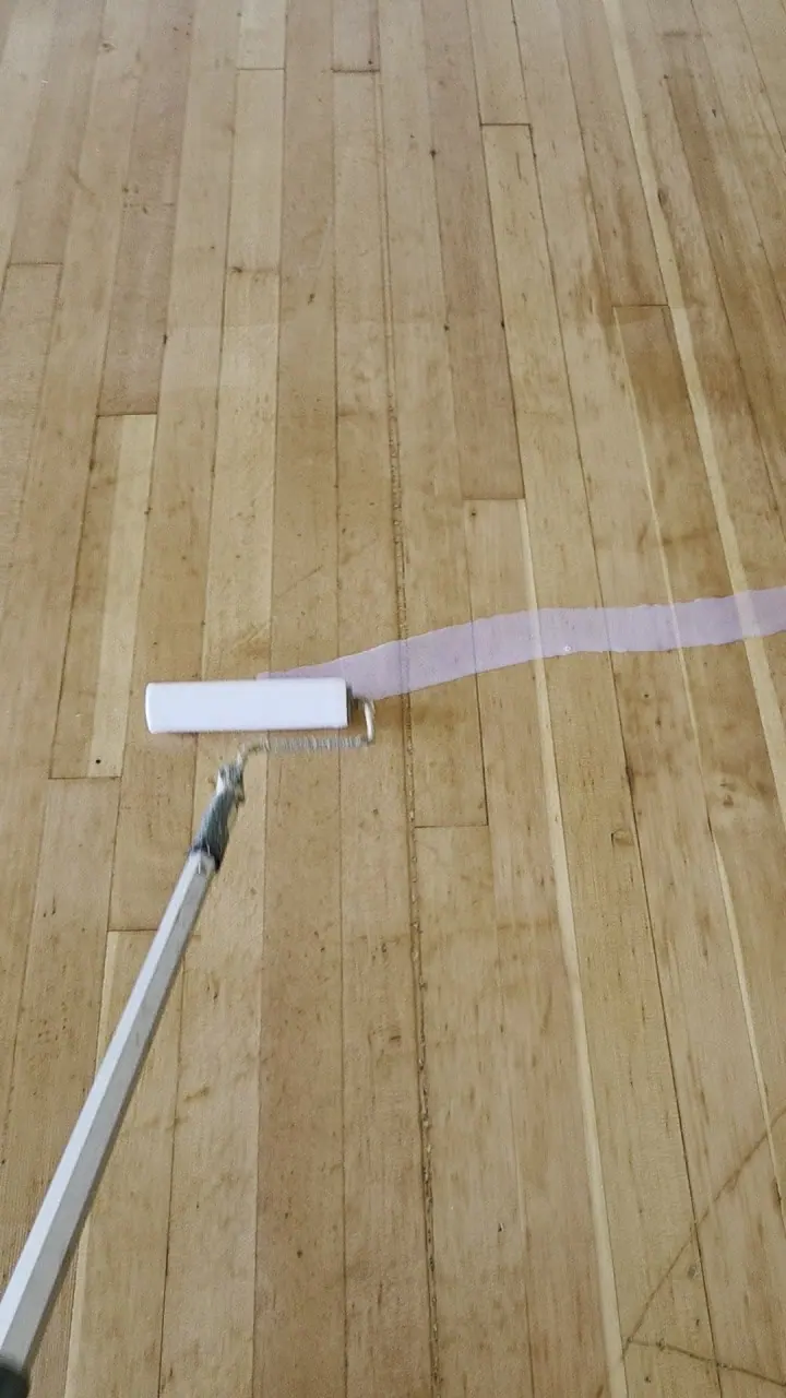 A roller applying a fresh coat of finish to a hardwood floor beside a white baseboard lined with blue tape.